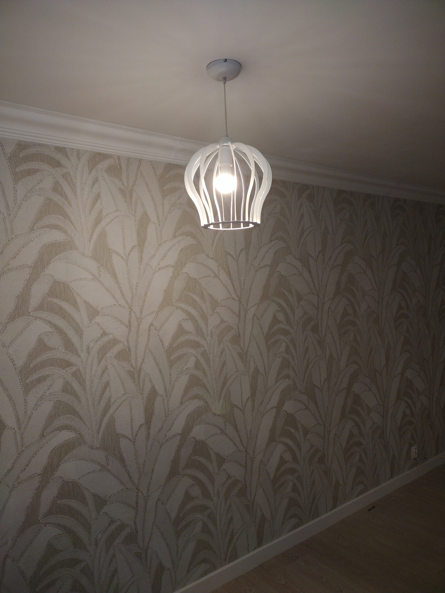 Nowoczesna lampa wisząca z białym ażurowym kloszem, oświetlająca ścianę pokrytą tapetą w motywy roślinne, z widoczną listwą przypodłogową i fragmentem parkietu.