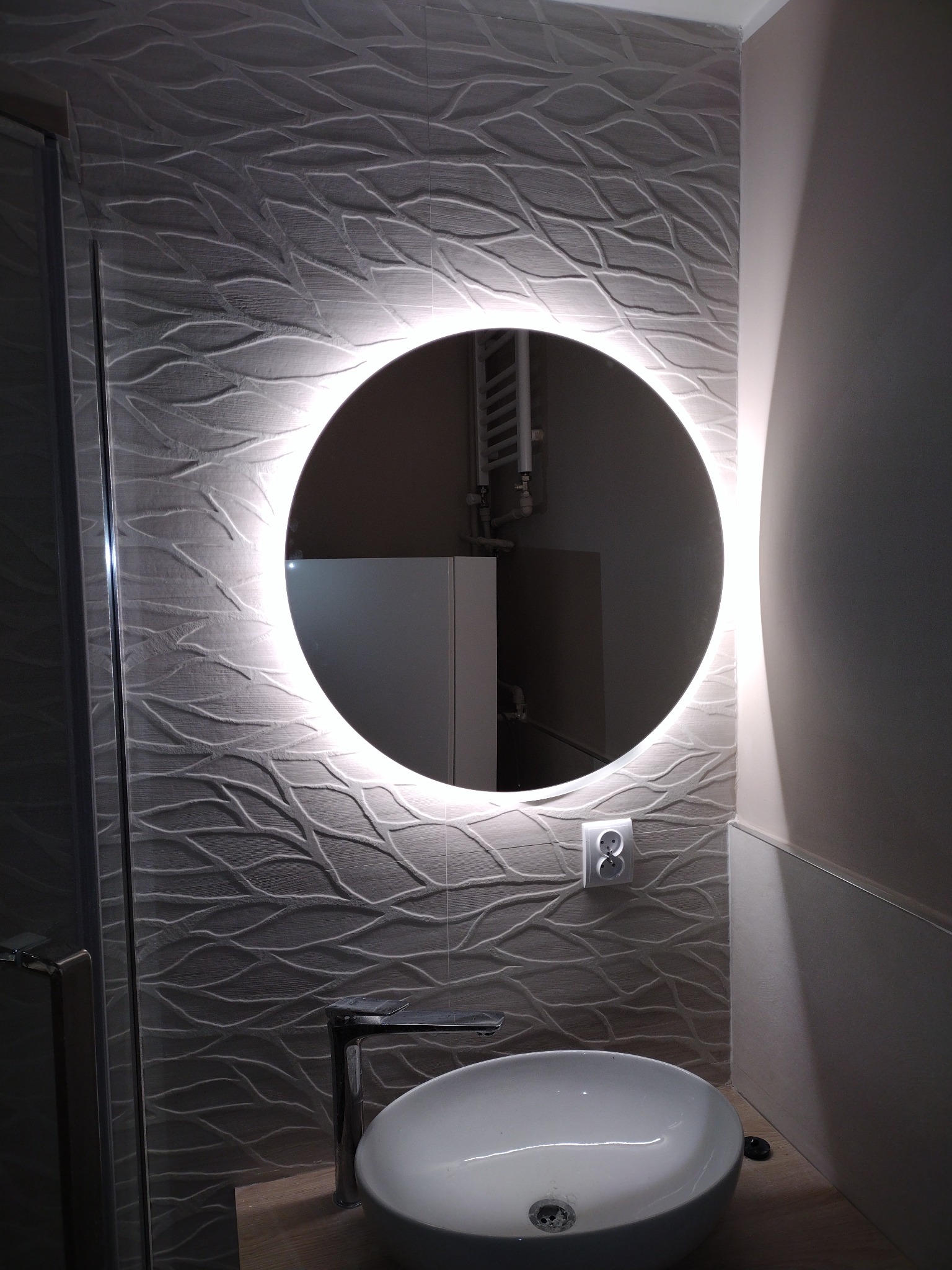 Nowoczesna łazienka z okrągłym lustrem LED, umywalką nablatową i płytkami ściennymi z subtelnym wzorem, widoczny fragment kabiny prysznicowej.