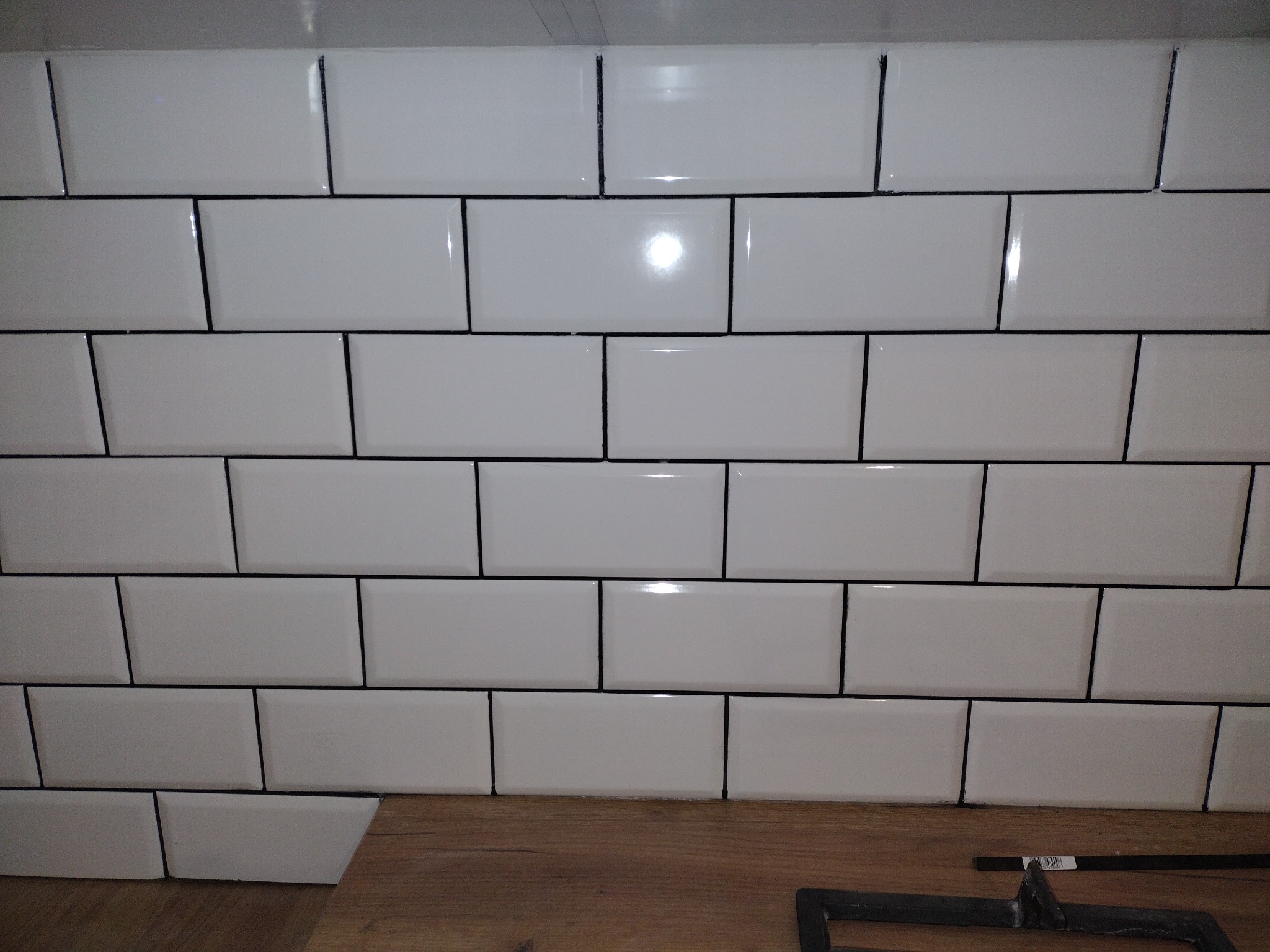 Świeżo ułożone białe, prostokątne płytki z ciemną fugą na ścianie, widoczny fragment drewnopodobnej podłogi i narzędzia glazurnicze.