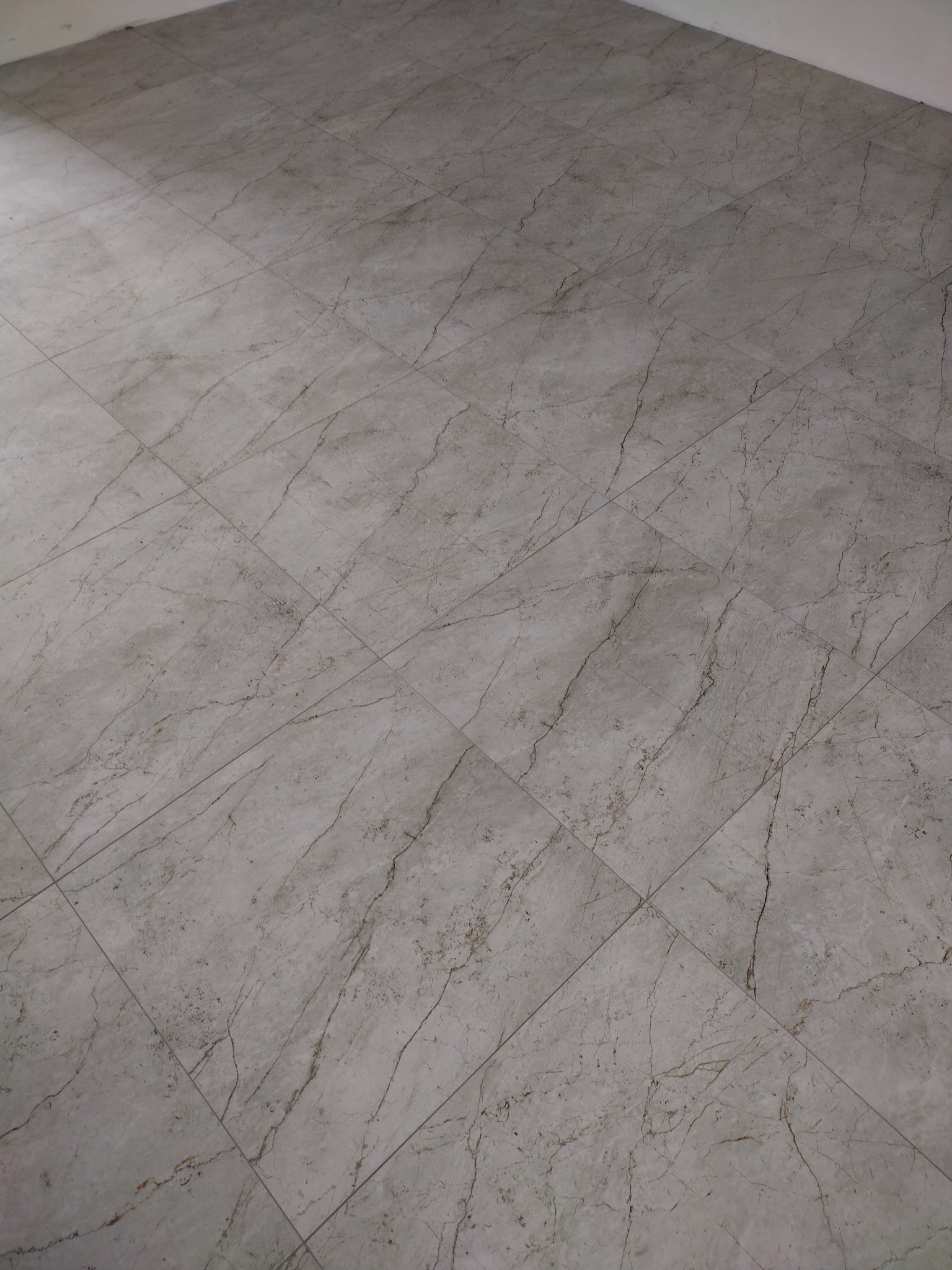 Jasnoszare płytki podłogowe imitujące marmur, ułożone ukośnie, z widocznymi fugami i delikatnym gradientem oświetlenia.