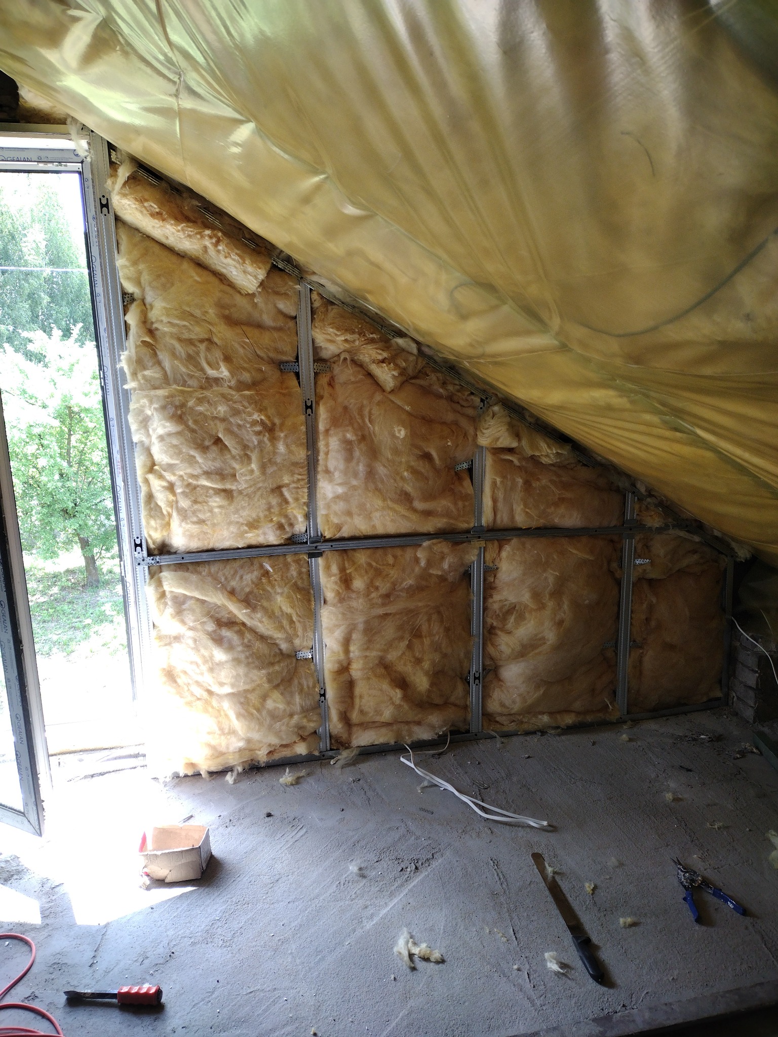 Izolacja poddasza wełną mineralną, widoczne profile metalowe i folia paroizolacyjna, narzędzia na podłodze, okno z widokiem na zieleń.