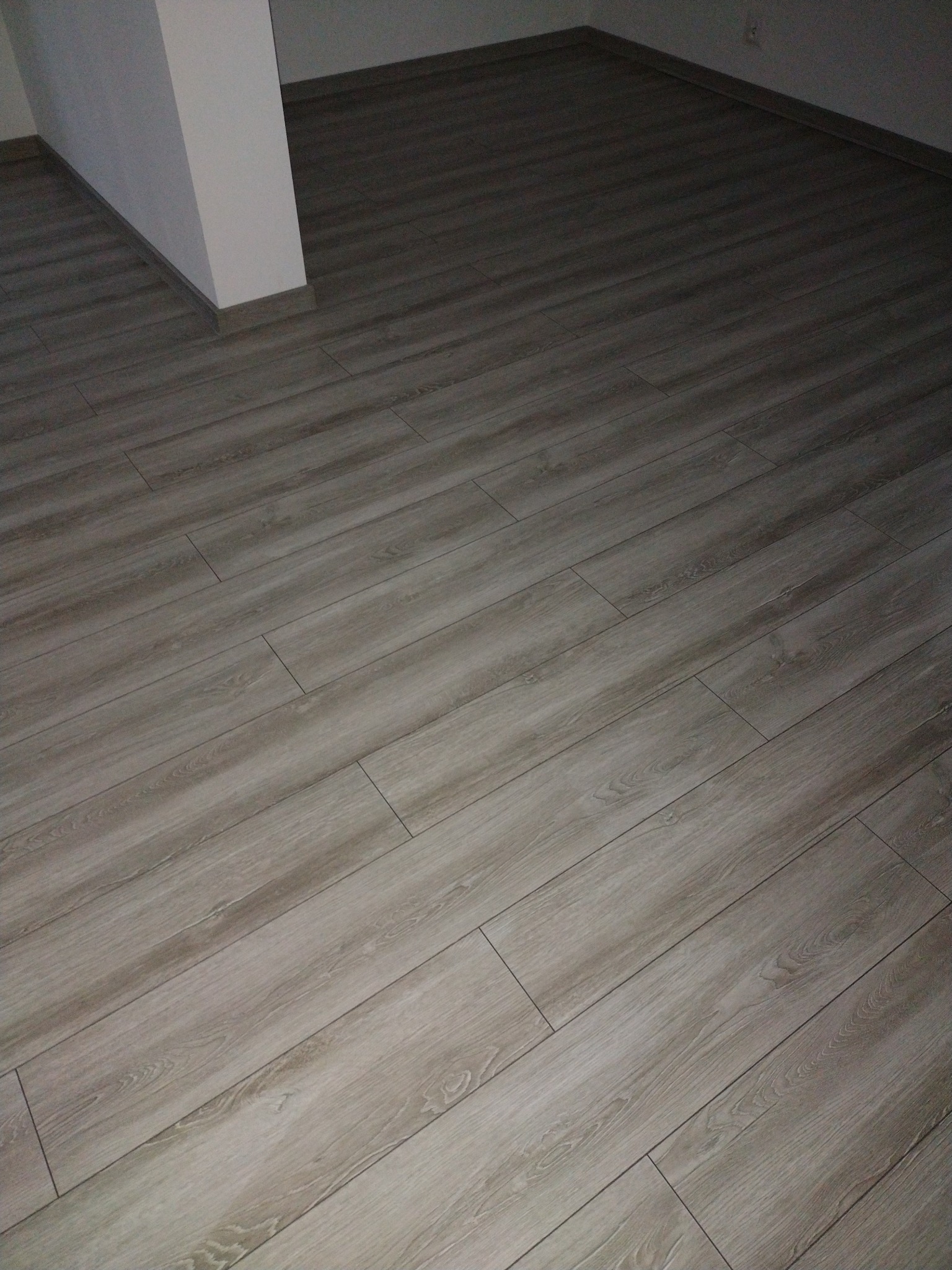 Ułożony parkiet w odcieniach szarości z widoczną listwą przypodłogową i fragmentem ściany w rogu pomieszczenia.