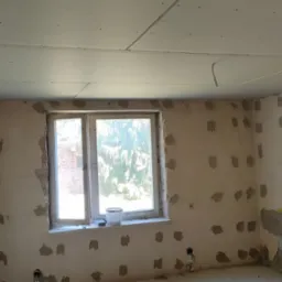 Pokój w trakcie remontu: zamontowana płyta karton gips na suficie, ściany z widocznymi miejscami po łataniu, okno z widokiem na zieleń, widoczne przewody elektryczne.