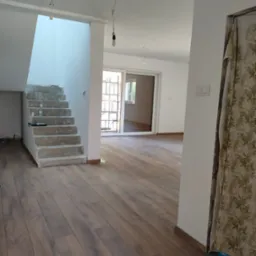 Wnętrze domu w trakcie remontu: surowe schody betonowe z widocznymi narzędziami i butelką, drewniana podłoga, białe ściany z wiszącymi żarówkami i zasłonięte drzwi w tle.