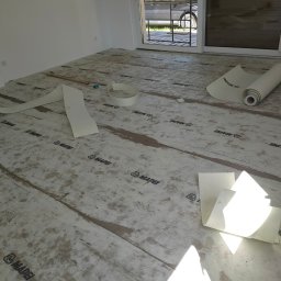 Budrem - Przygotowanie podłoża pod parkiet: rozłożona mata izolacyjna z widocznymi łączeniami i logotypami producenta, częściowo rozwinięta rolka maty, drewniana listwa i miarka na podłodze, okno balkonowe...