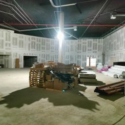Budrem - Wnętrze w trakcie prac wykończeniowych: ściany pokryte płytami gipsowo-kartonowymi z widocznymi łączeniami, paleta z materiałami budowlanymi na pierwszym planie, instalacje pod sufitem, widoczny...