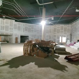 Budrem - Wnętrze w trakcie prac wykończeniowych: ściany z płyt karton-gips, paleta z materiałami budowlanymi na betonowej podłodze, widoczne instalacje pod sufitem.