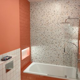 Budrem - Nowoczesna łazienka z wanną obudowaną pomarańczowymi płytkami, mozaikową ścianą, szklanym parawanem nawannowym i chromowaną armaturą, uzupełniona o białe płytki i detale w odcieniach pomarańczy.
