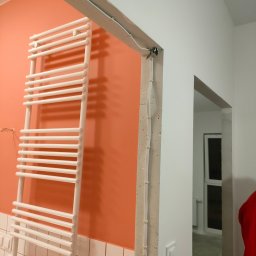 Budrem - Biały grzejnik drabinkowy na pomarańczowej ścianie z białymi płytkami w dolnej części, widoczne kable przymocowane do surowej ściany oraz przejście do innego pomieszczenia.