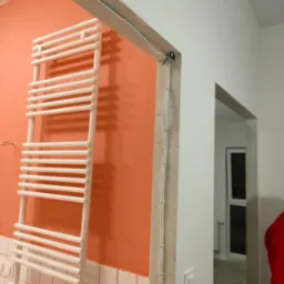 Biały grzejnik drabinkowy na pomarańczowej ścianie z białymi płytkami w dolnej części, widoczne kable przymocowane do surowej ściany oraz przejście do innego pomieszczenia.