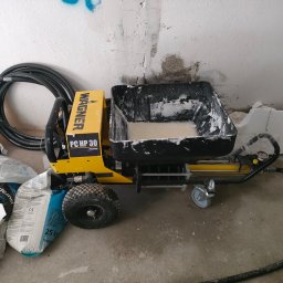 Budrem - Żółta maszyna Wagner PC HP 30 do natrysku gładzi z czarnym zbiornikiem wypełnionym białą masą, obok worki z zaprawą, kabel i fragment ściany w trakcie prac wykończeniowych.
