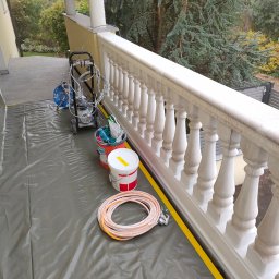 Budrem - Przygotowanie do malowania balustrady na balkonie: rozłożona folia ochronna, taśma malarska, agregat malarski, wiadro z farbą i akcesoriami malarskimi.