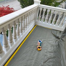 Budrem - Biała balustrada balkonu zabezpieczona folią malarską i żółtą taśmą maskującą przed malowaniem, widoczne dwie rolki taśmy malarskiej.