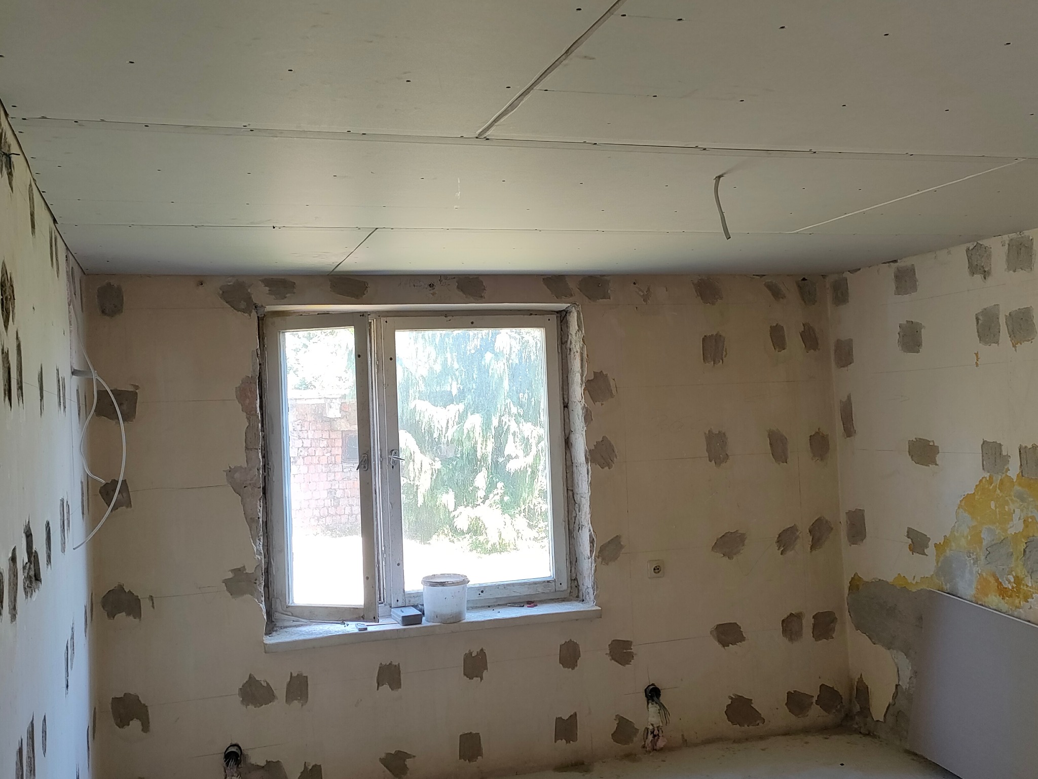 Pokój w trakcie remontu: zamontowana płyta karton gips na suficie, ściany z widocznymi miejscami po łataniu, okno z widokiem na zieleń, widoczne przewody elektryczne.