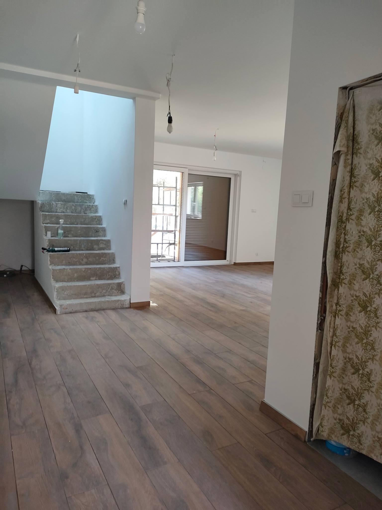 Wnętrze domu w trakcie remontu: surowe schody betonowe z widocznymi narzędziami i butelką, drewniana podłoga, białe ściany z wiszącymi żarówkami i zasłonięte drzwi w tle.