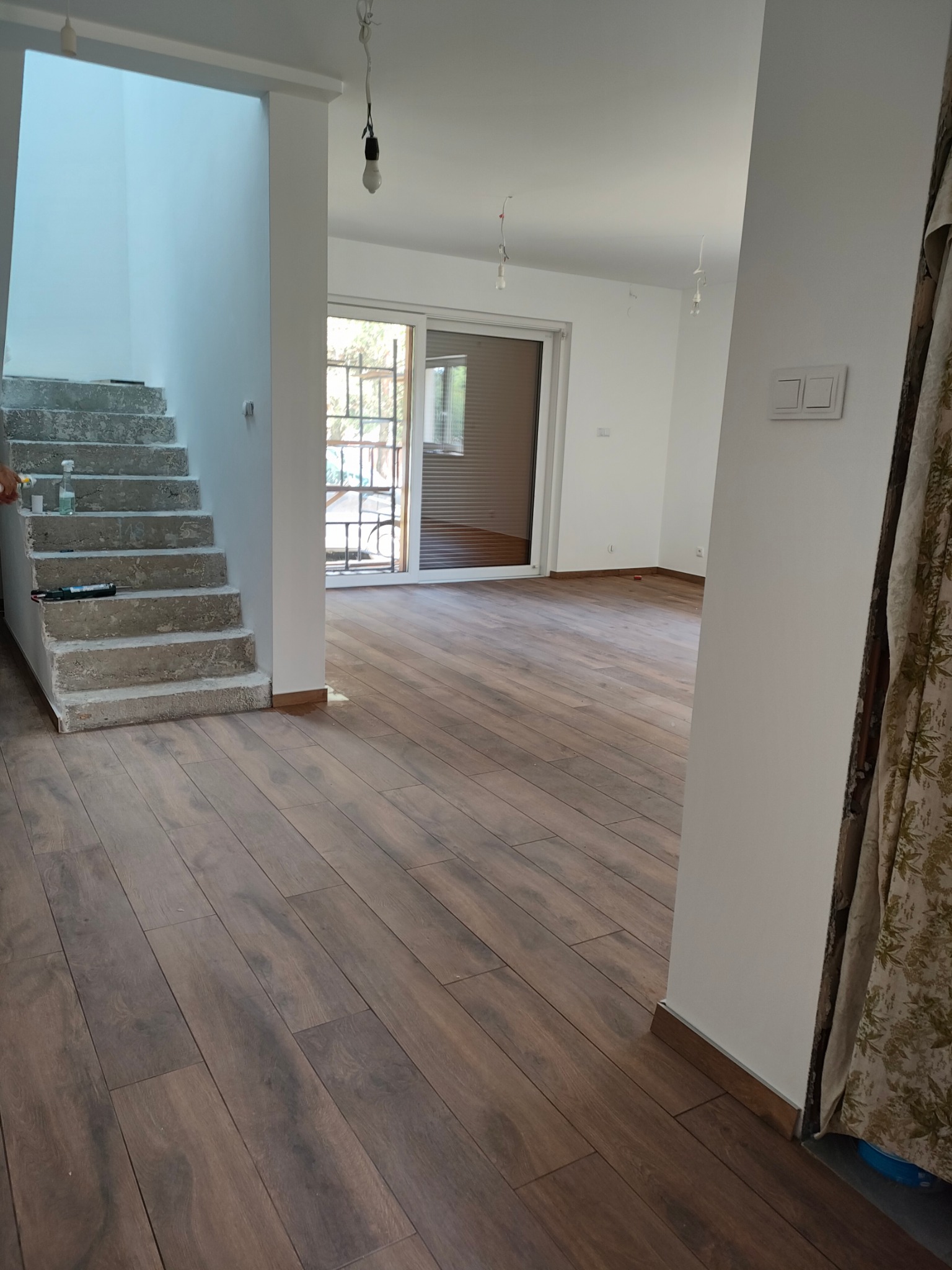 Układanie paneli podłogowych w nowym domu, widoczne surowe schody betonowe i okno balkonowe z żaluzjami.