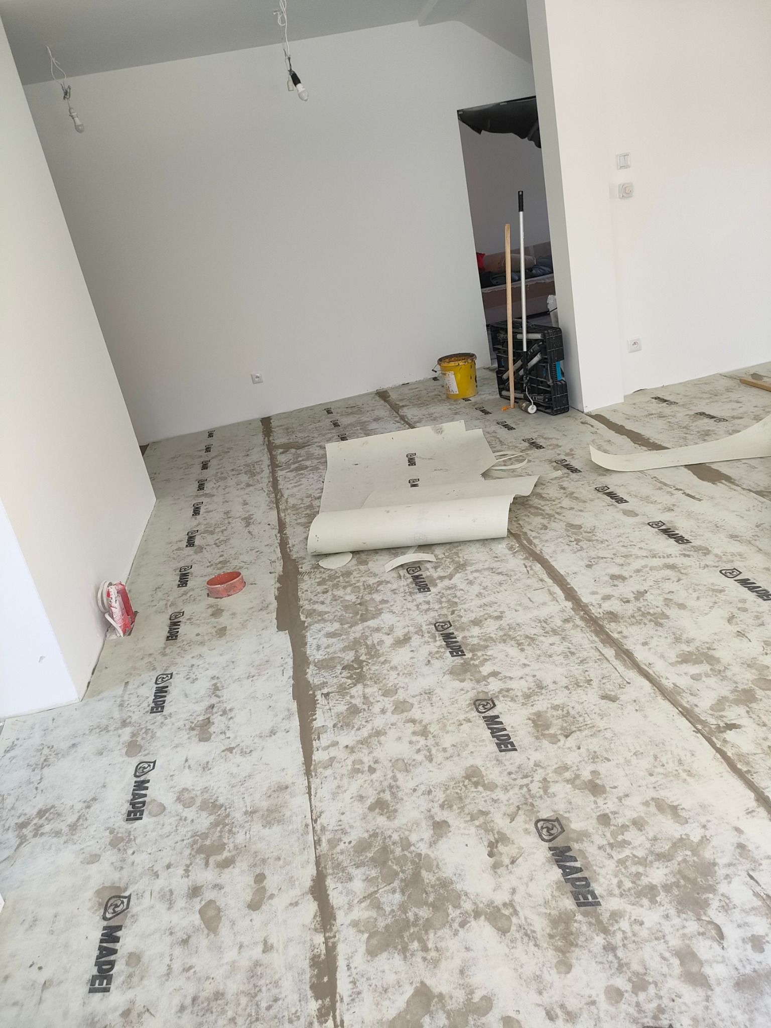 Przygotowanie podłoża pod parkiet: gruntowanie wylewki samopoziomującej z widocznymi wałkami folii ochronnej z nadrukiem MAPEI, wiadro i narzędzia w tle, surowy stan wnętrza.