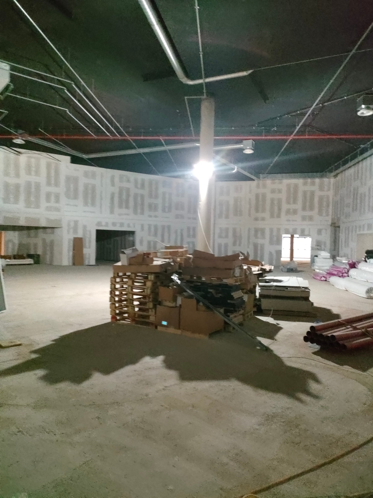 Wnętrze w trakcie prac wykończeniowych: ściany z płyt karton-gips, paleta z materiałami budowlanymi na betonowej podłodze, widoczne instalacje pod sufitem.