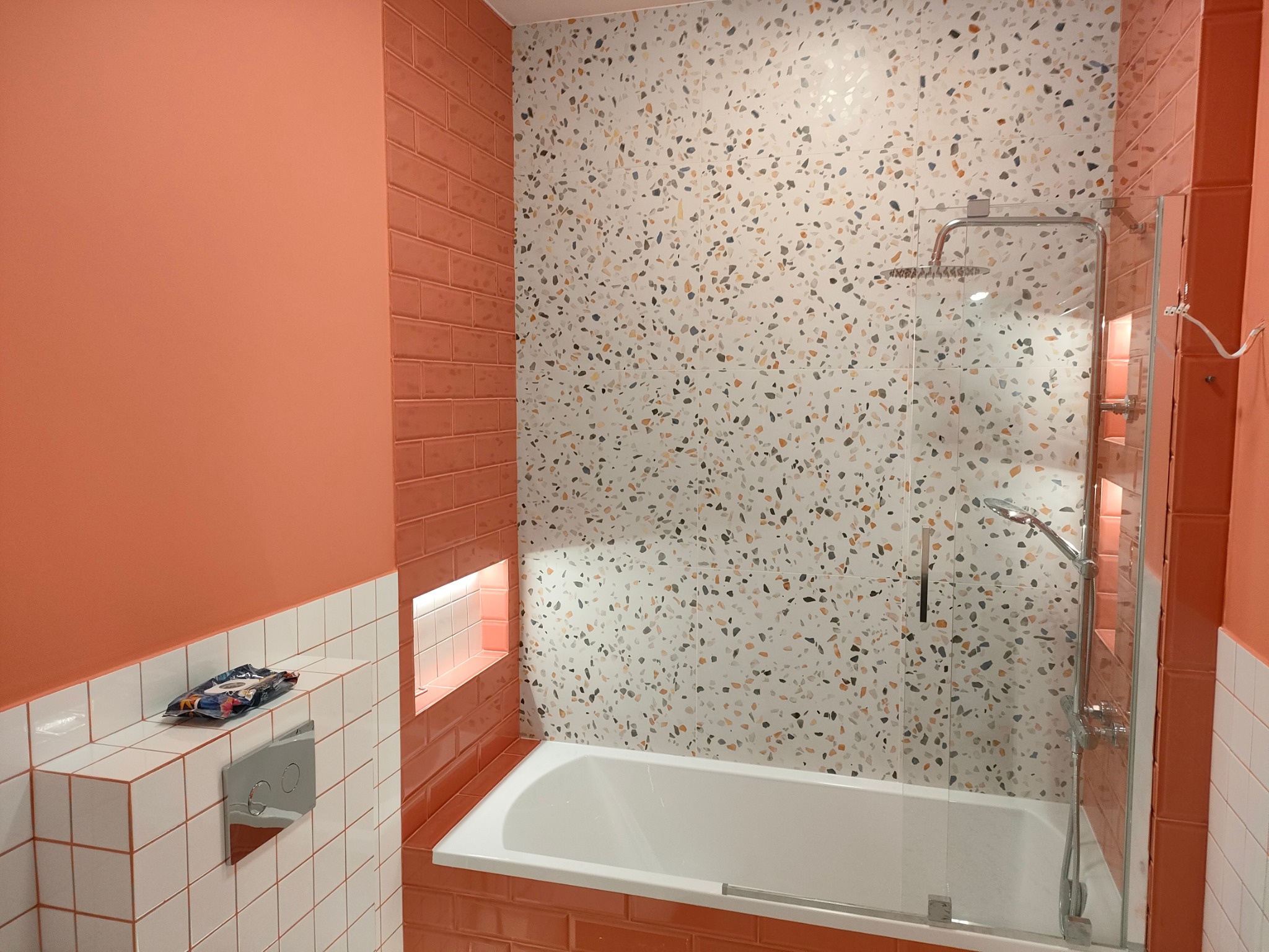 Nowoczesna łazienka z wanną obudowaną pomarańczowymi płytkami, mozaikową ścianą, szklanym parawanem nawannowym i chromowaną armaturą, uzupełniona o białe płytki i detale w odcieniach pomarańczy.