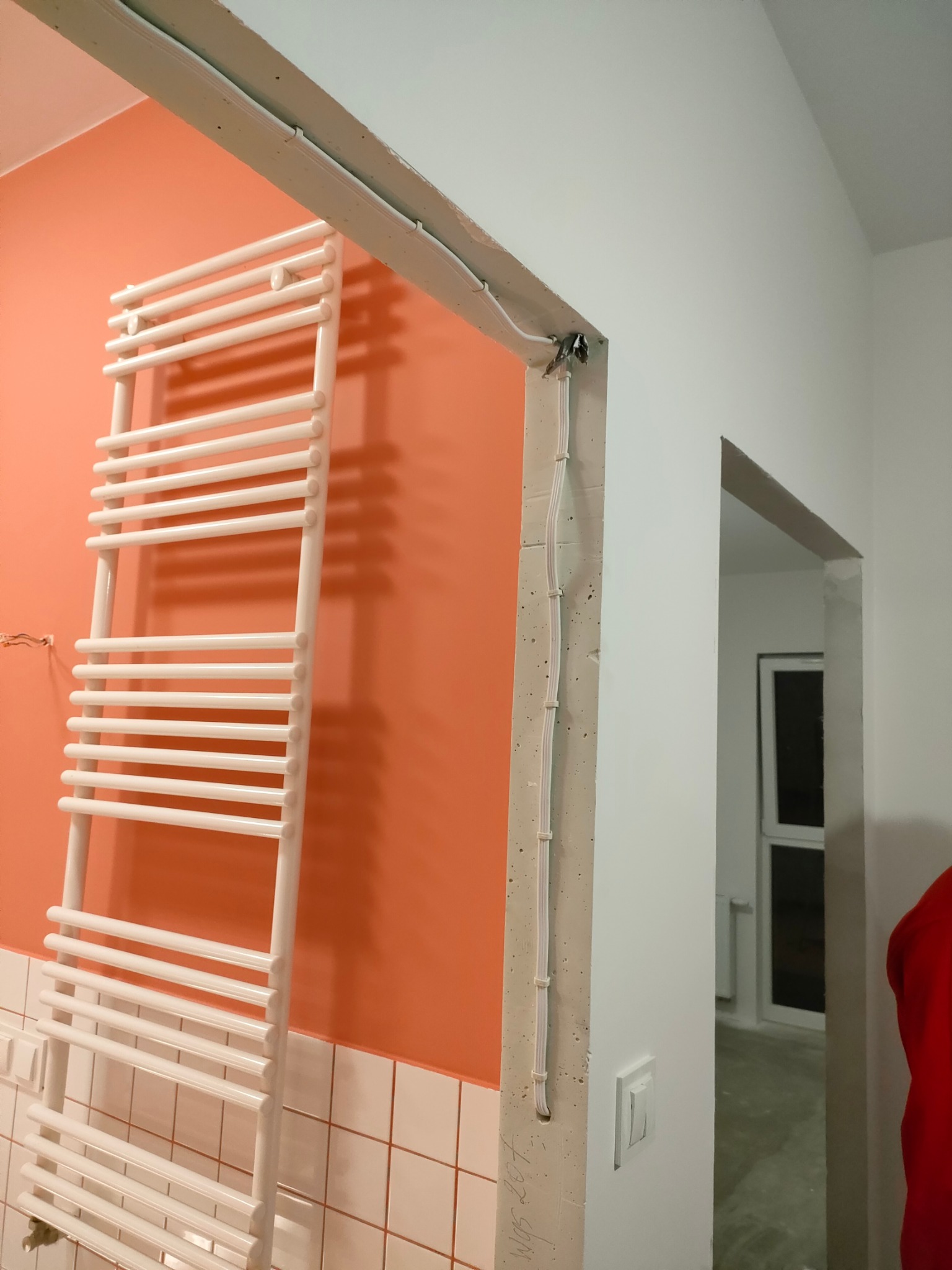 Biały grzejnik drabinkowy na pomarańczowej ścianie z białymi płytkami w dolnej części, widoczne kable przymocowane do surowej ściany oraz przejście do innego pomieszczenia.
