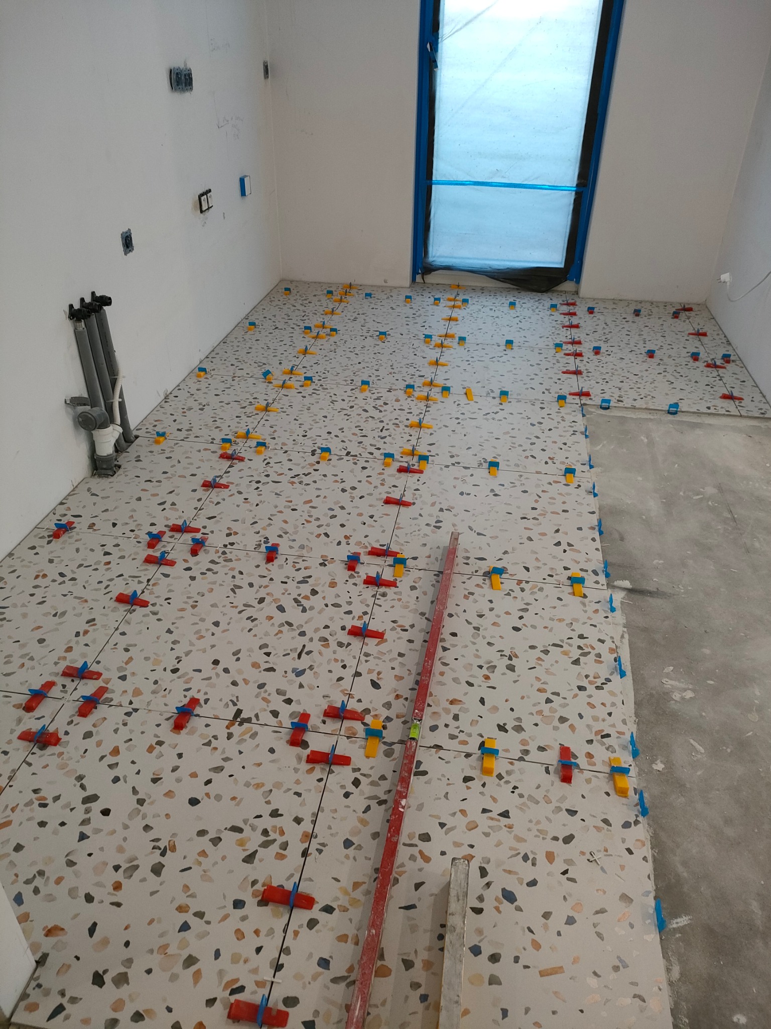 Układanie terakoty w łazience: podłoga z płytek w stylu lastryko z systemem poziomowania, widoczne piony kanalizacyjne przy ścianie, okno zabezpieczone folią.