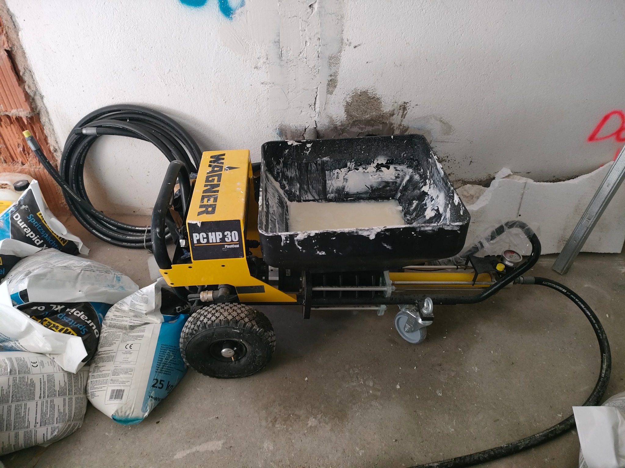 Żółta maszyna Wagner PC HP 30 do natrysku gładzi z czarnym zbiornikiem wypełnionym białą masą, obok worki z zaprawą, kabel i fragment ściany w trakcie prac wykończeniowych.