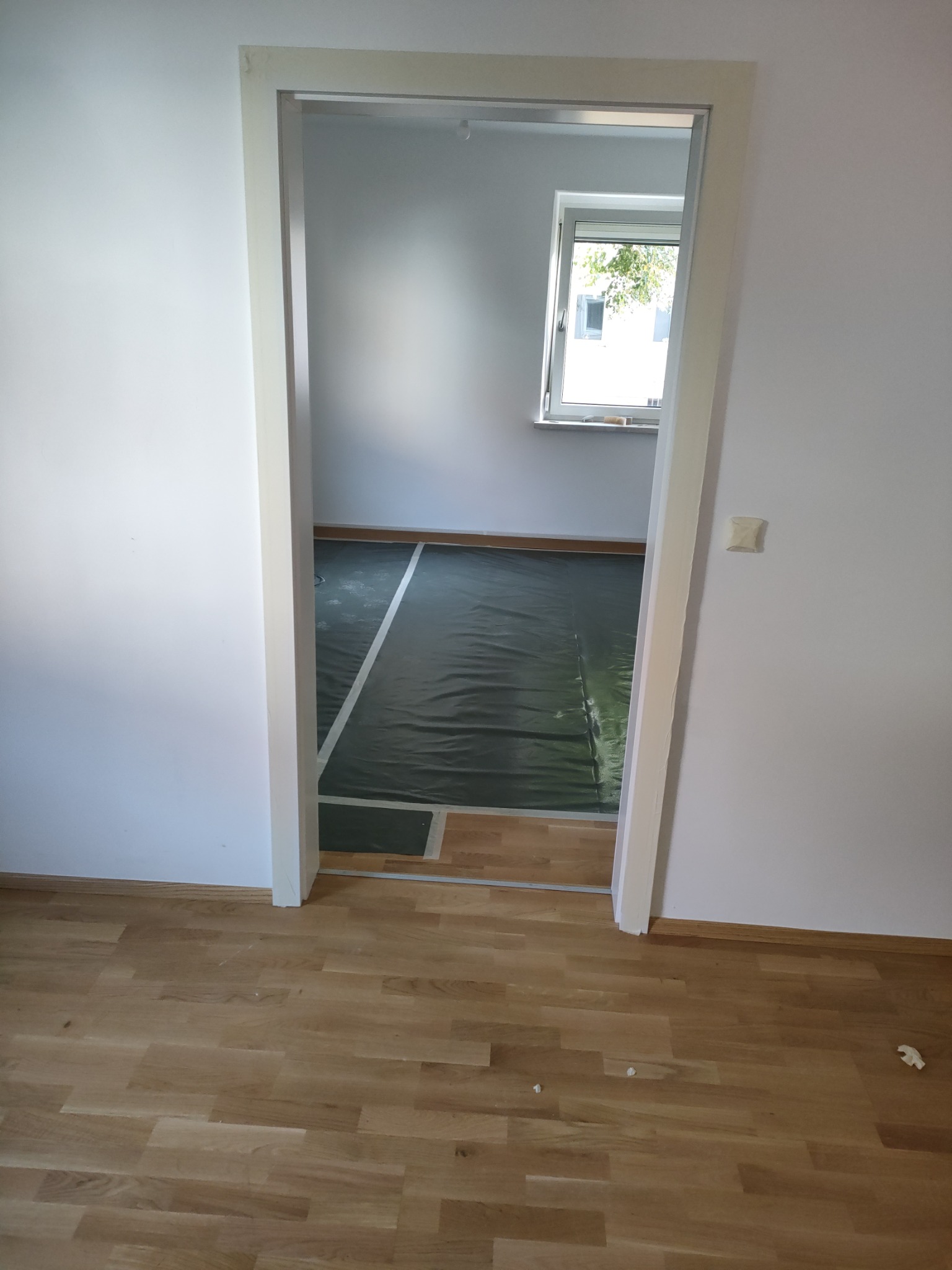 Widok przez białe futryny na pokój z zabezpieczoną folią podłogą, świeżo ułożony parkiet na pierwszym planie, okno w tle.