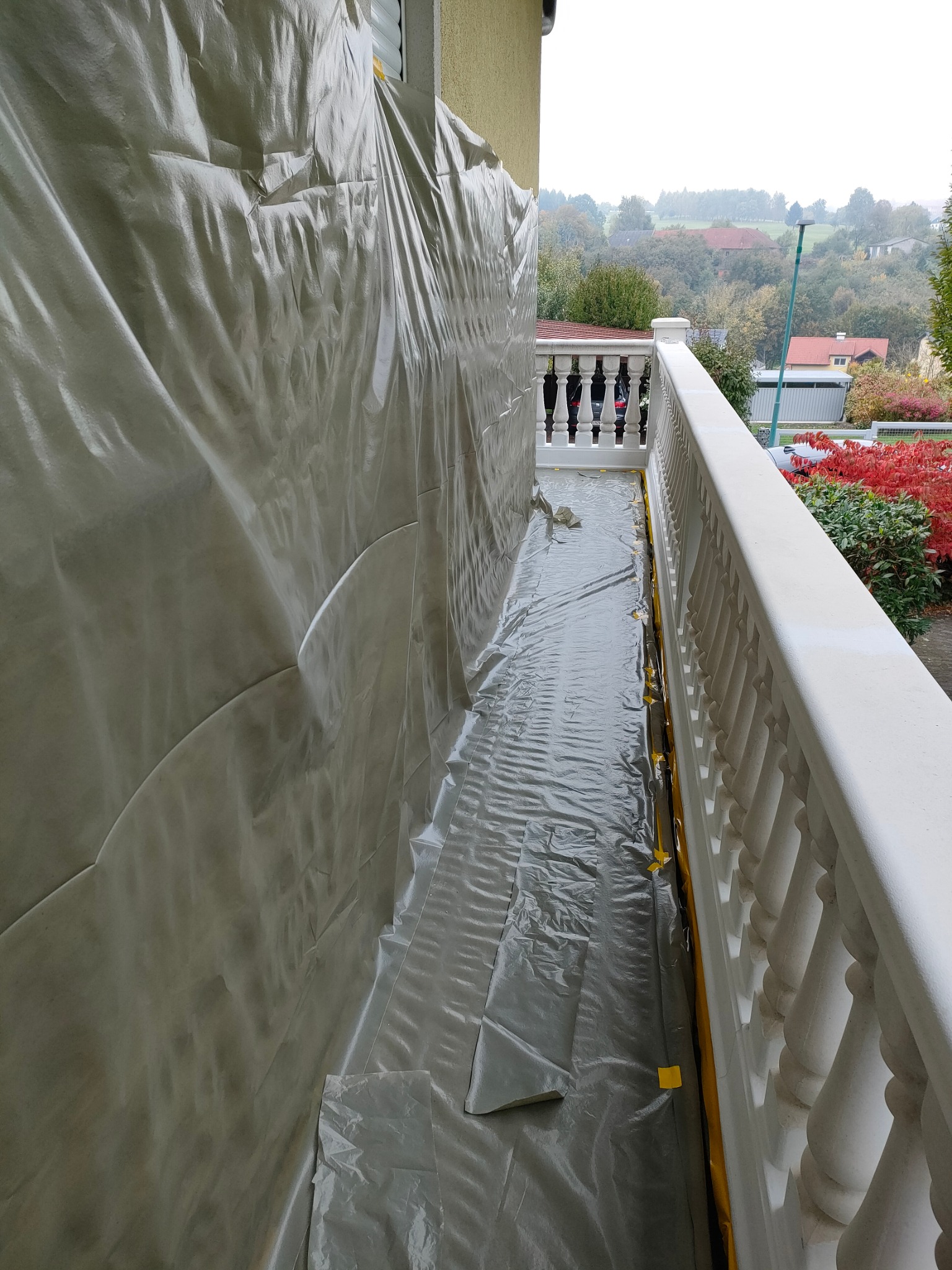 Balkon zabezpieczony folią malarską podczas prac remontowych, widoczne białe balustrady i otoczenie budynku.