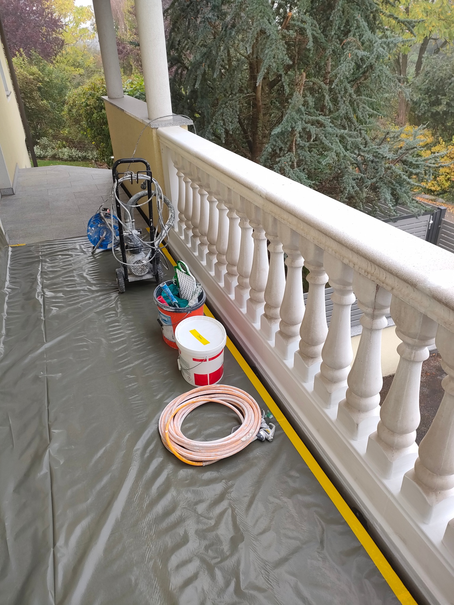 Przygotowanie do malowania balustrady na balkonie: rozłożona folia ochronna, taśma malarska, agregat malarski, wiadro z farbą i akcesoriami malarskimi.