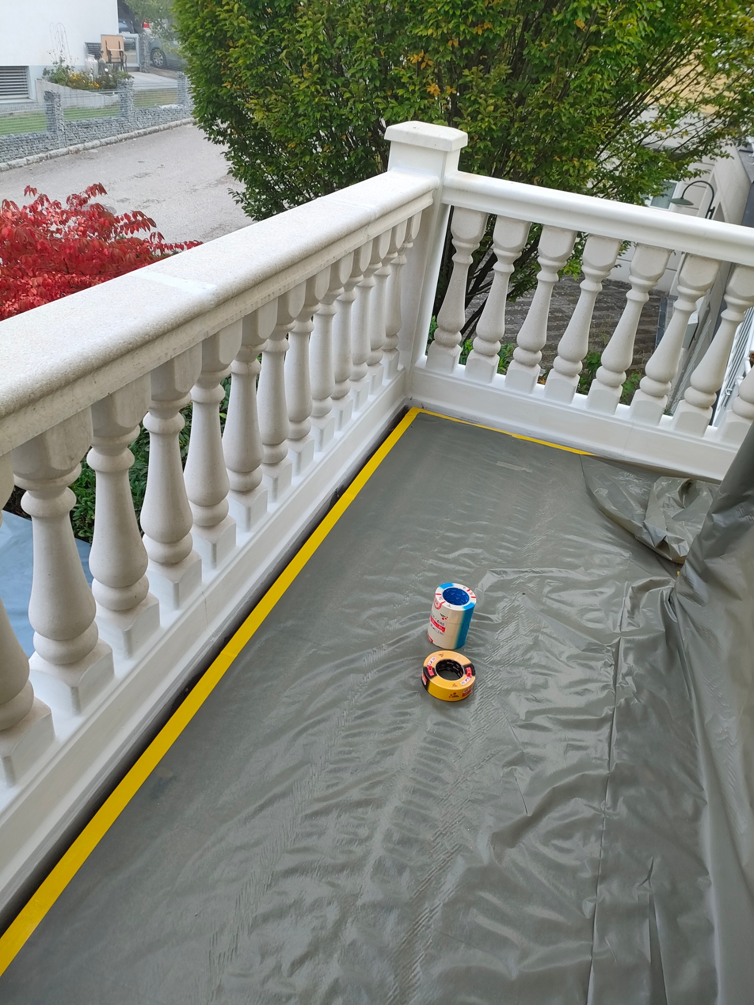 Biała balustrada balkonu zabezpieczona folią malarską i żółtą taśmą maskującą przed malowaniem, widoczne dwie rolki taśmy malarskiej.