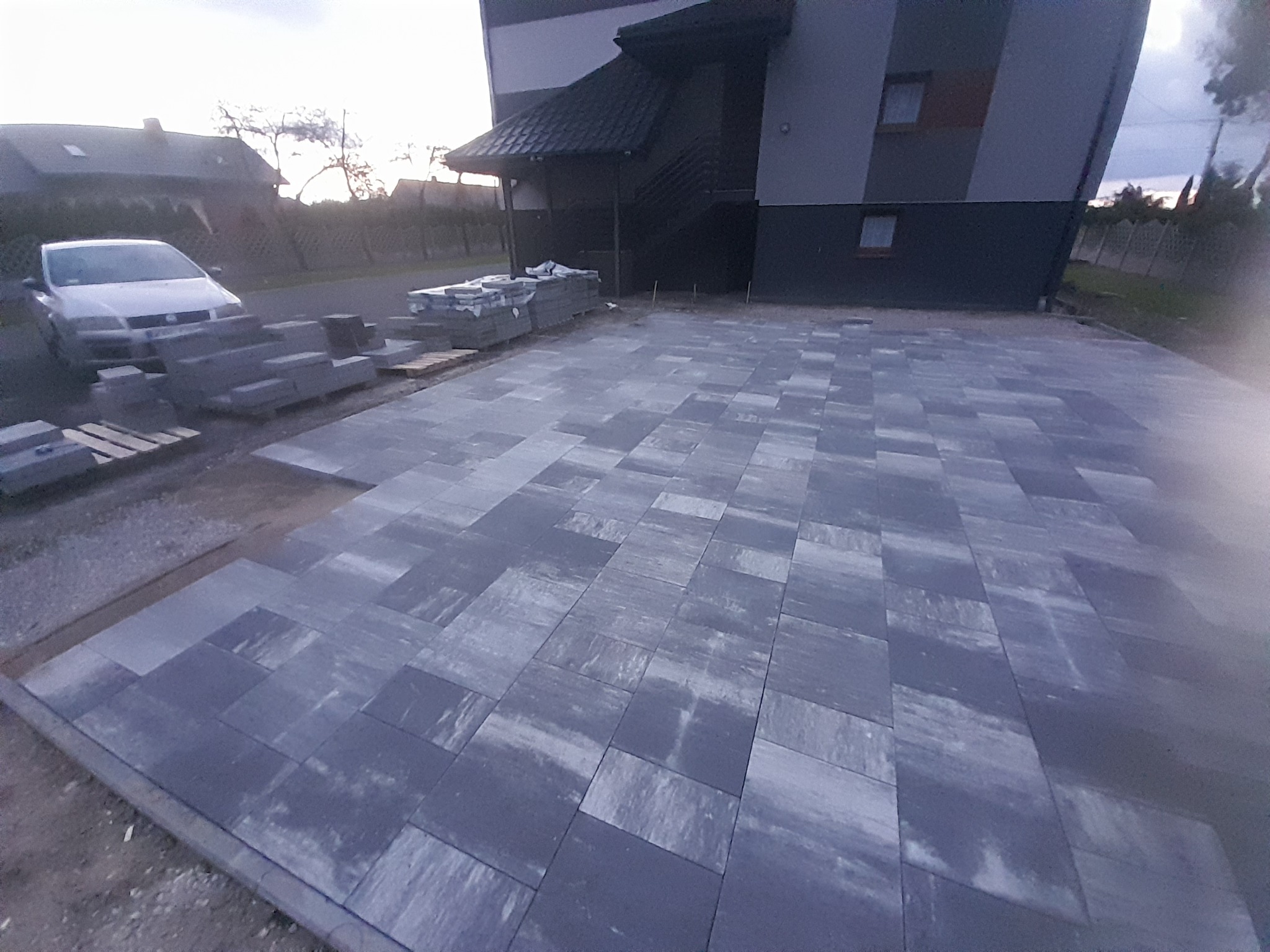 Ułożona szara kostka brukowa na podjeździe przed nowoczesnym budynkiem, palety z kostką w tle, widoczny samochód osobowy.