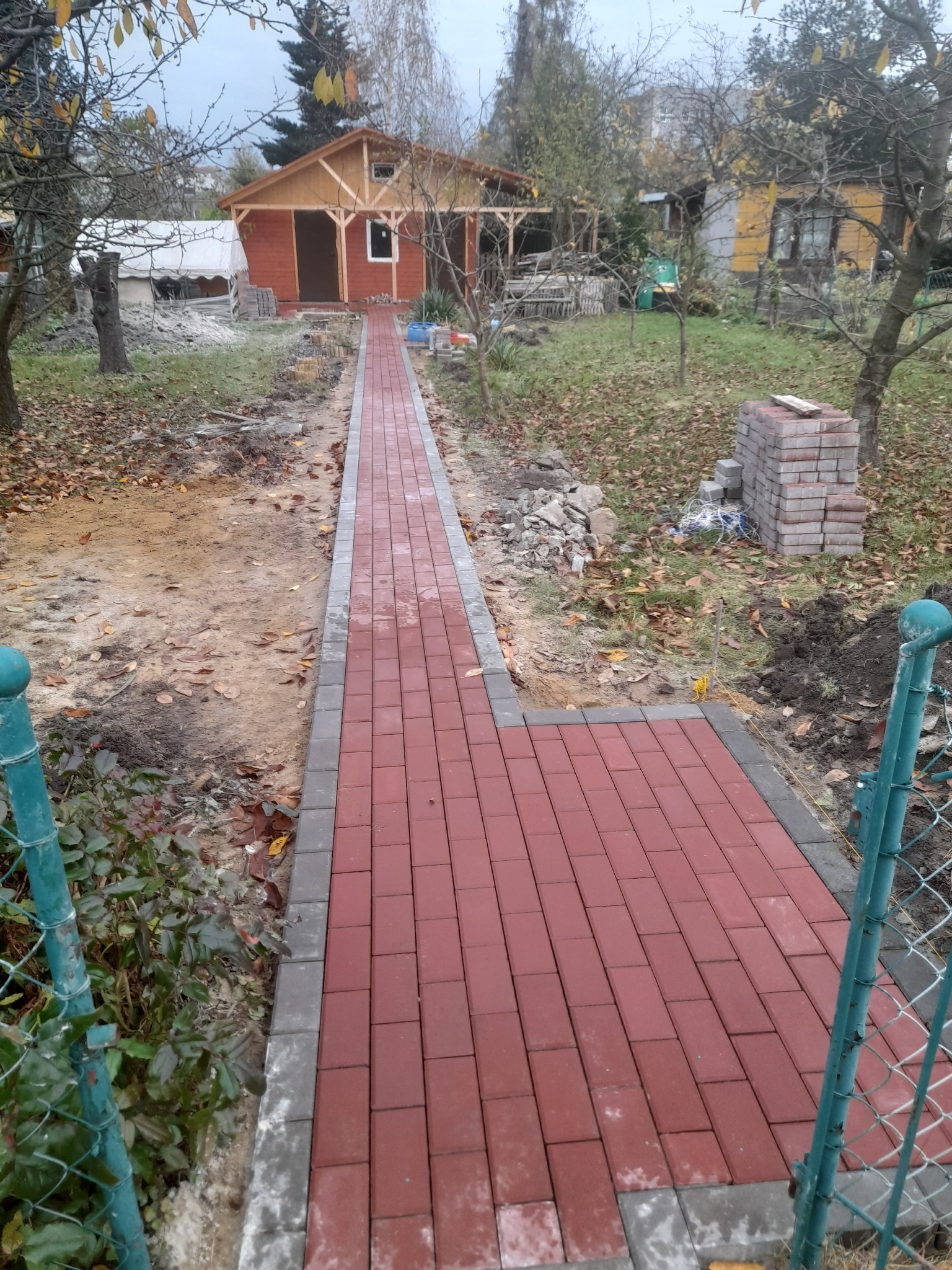 Czerwona kostka brukowa z szarymi obrzeżami prowadząca do drewnianego domku w ogrodzie, widoczne niedokończone prace i sterta kostki obok ścieżki.