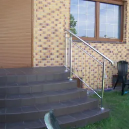 Zewnętrzne schody wejściowe z ciemnobrązowych płytek z chromowaną balustradą, widok na elewację z cegły klinkierowej i okno z kratownicą.