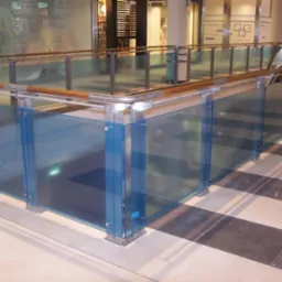 Szklana balustrada z drewnianym poręczą w przestrzeni komercyjnej, widok na schody ruchome i sklepy w tle.