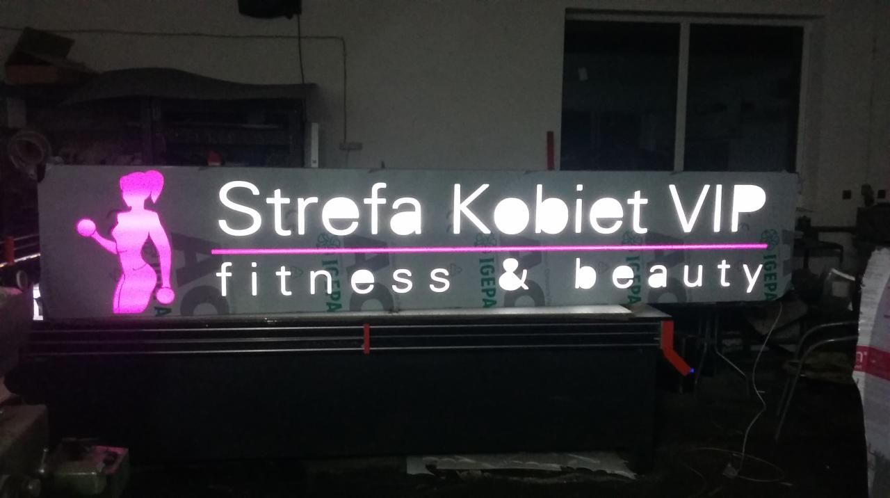 Podświetlany szyld reklamowy 'Strefa Kobiet VIP fitness & beauty' z różową sylwetką kobiety ćwiczącej z hantlami, wykonany z materiałów kompozytowych.