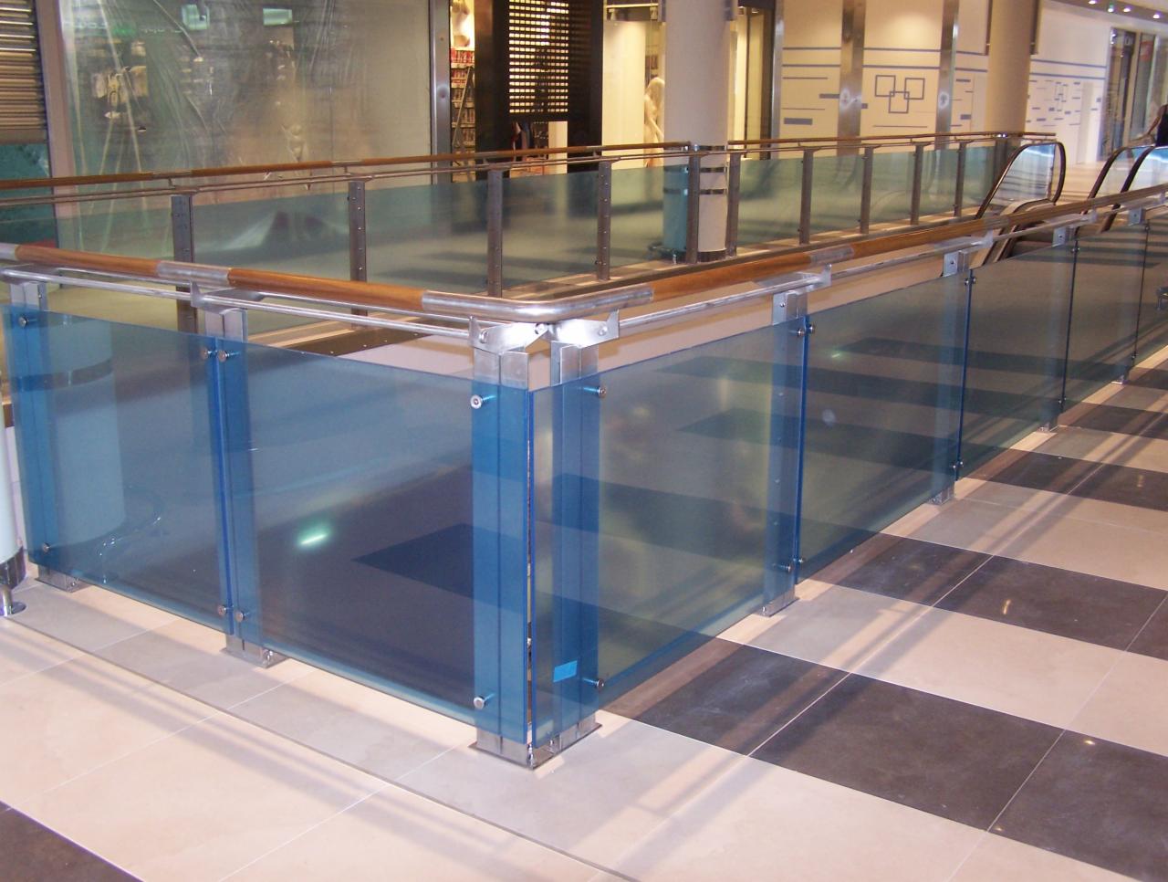 Szklana balustrada z drewnianym poręczą w przestrzeni komercyjnej, widok na schody ruchome i sklepy w tle.