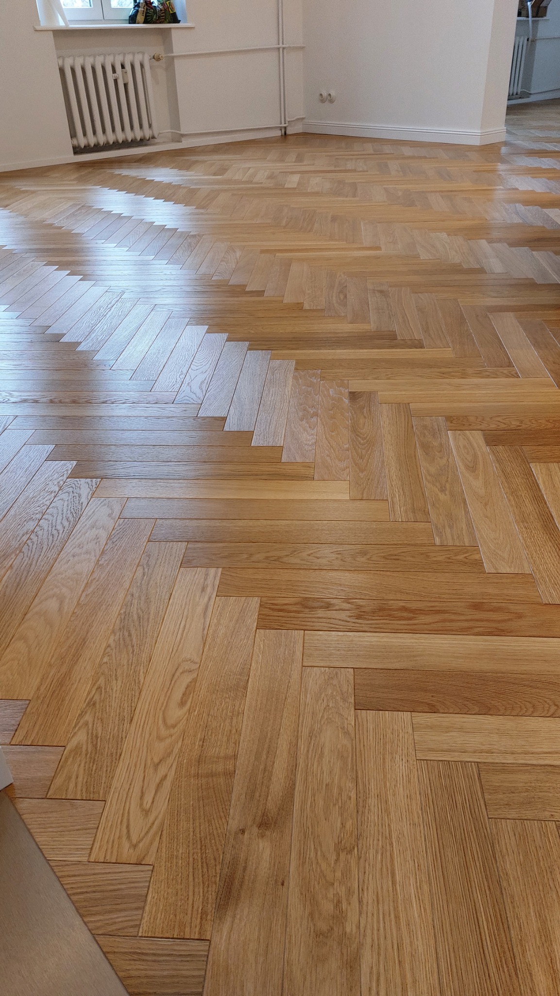 Ułożony parkiet w jodełkę francuską, jasne drewno, widoczne grzejniki i białe ściany w tle.