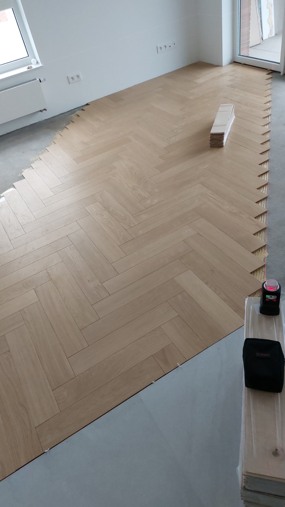 Układanie jasnego parkietu w jodełkę francuską, widoczne narzędzia: poziomica laserowa i torba na narzędzia, oraz stos nieużytych desek parkietowych.