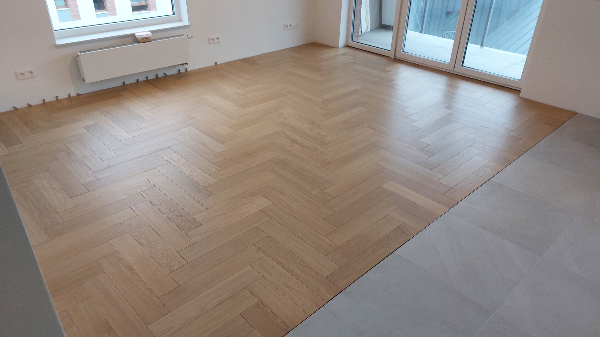 Jasny parkiet ułożony we wzór jodełki francuskiej, widoczne listwy dystansowe przy ścianie, fragment podłogi wyłożonej szarymi płytkami, okno balkonowe w tle.