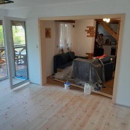 MIWA EKO - Jasna, drewniana podłoga po renowacji w przestronnym wnętrzu z widokiem na taras. Częściowo zakryte meble sugerują trwające prace wykończeniowe.