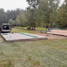 MIWA EKO - Przygotowanie do budowy tarasu drewnianego: betonowa podstawa, deski, piła stołowa, samochód dostawczy i osoba w tle, na tle drzew.