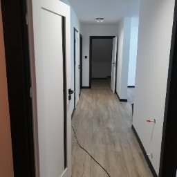 MIWA EKO - Wykończony korytarz z panelami podłogowymi w odcieniach szarości i białymi drzwiami z czarnymi ościeżnicami. Widoczne detale wykończeniowe i oświetlenie sufitowe.