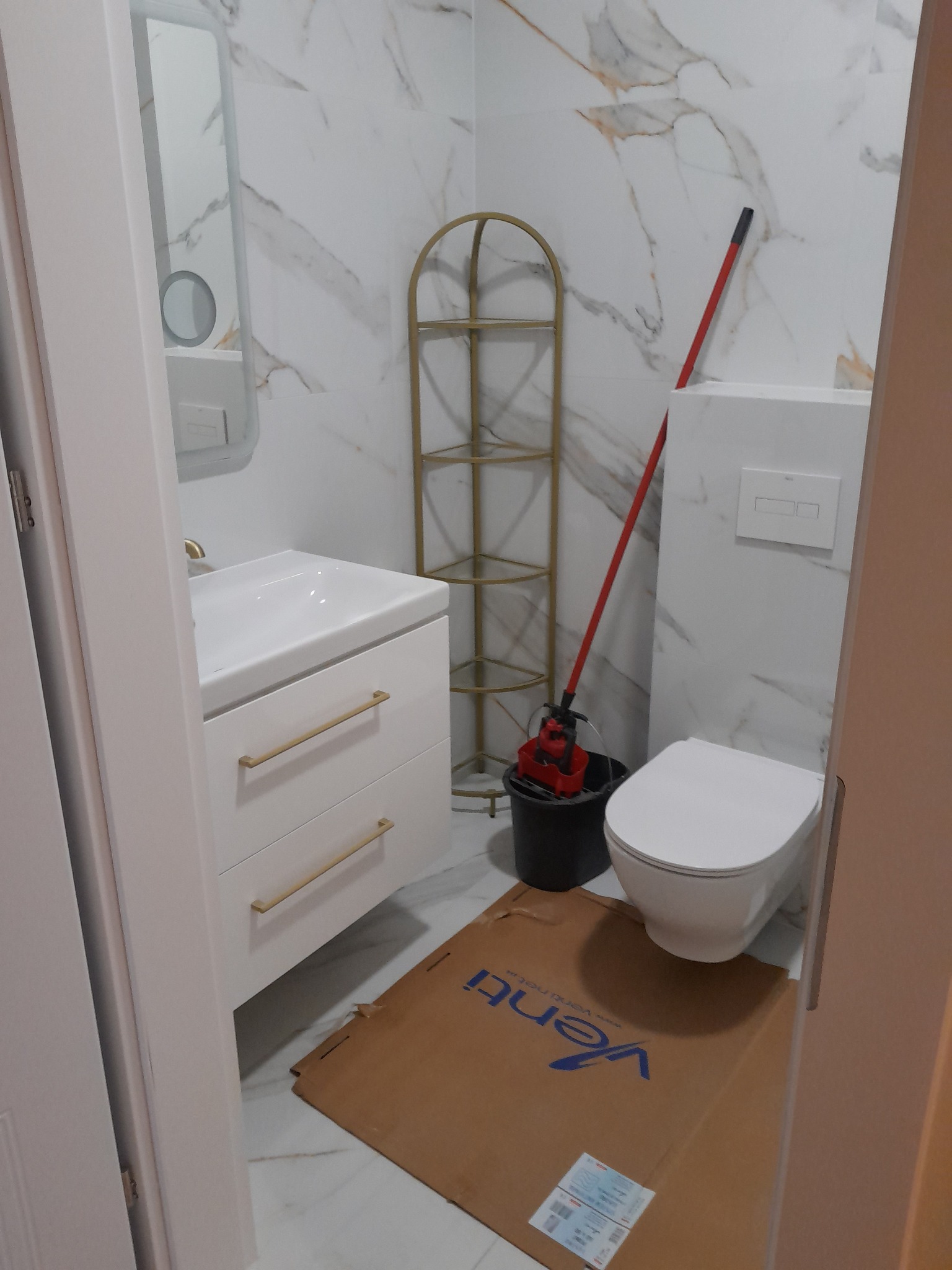 Biała łazienka z marmurowymi płytkami, złotymi akcentami i nowoczesną armaturą. Widoczny fragment podłogi zabezpieczony kartonem.