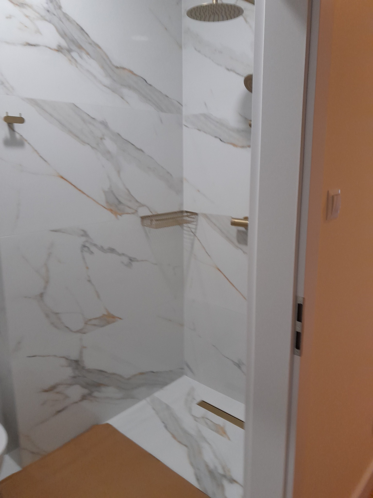 Kabina prysznicowa z białymi płytkami imitującymi marmur, złota armatura, odpływ liniowy. Widok zza uchylonych drzwi, fragment podłogi poza kabiną.