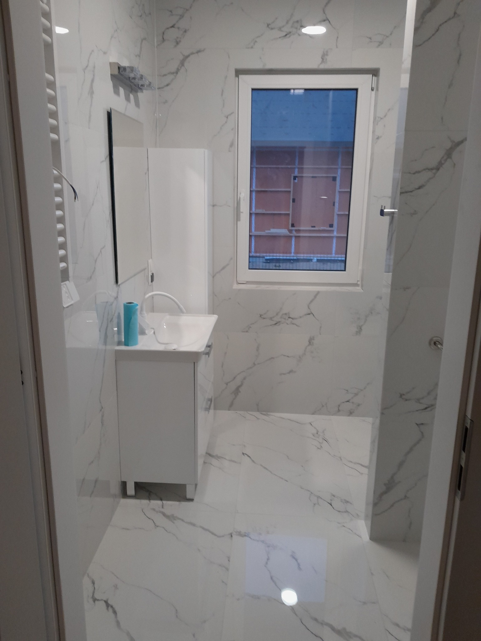 Wykończona łazienka w Przykory z białymi płytkami imitującymi marmur, białą szafką pod umywalką i oknem. Nowoczesny design i minimalistyczne dodatki.