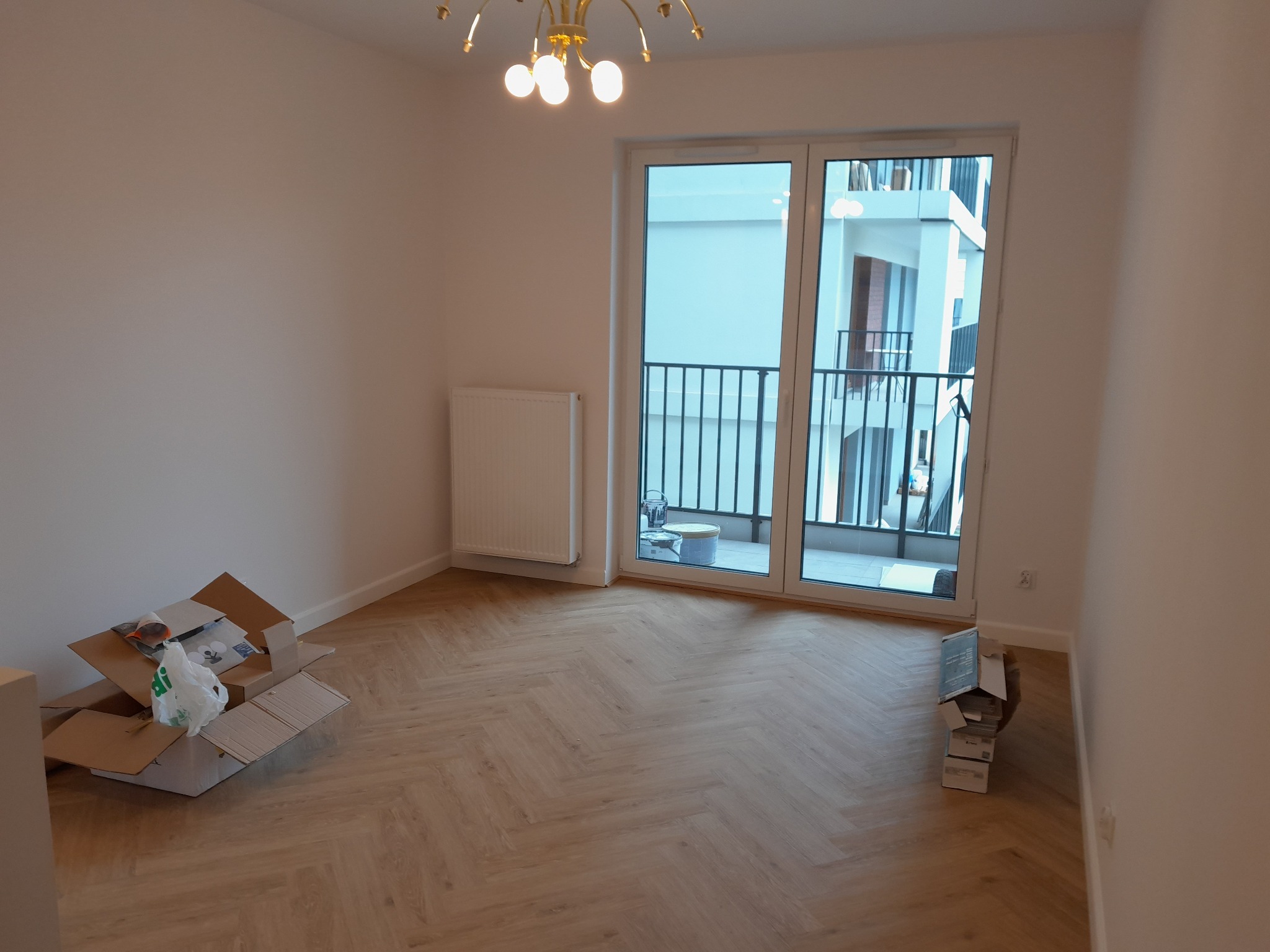 Jasne panele ułożone w jodełkę w pustym pokoju z balkonem. Widoczne kartony, grzejnik i złoty żyrandol. Nowe budownictwo, minimalistyczny styl.