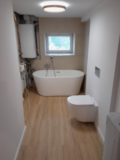 Nowoczesna łazienka po remoncie: wolnostojąca wanna, podwieszana toaleta, drewnopodobne panele podłogowe, beżowe ściany i okrągła lampa sufitowa.