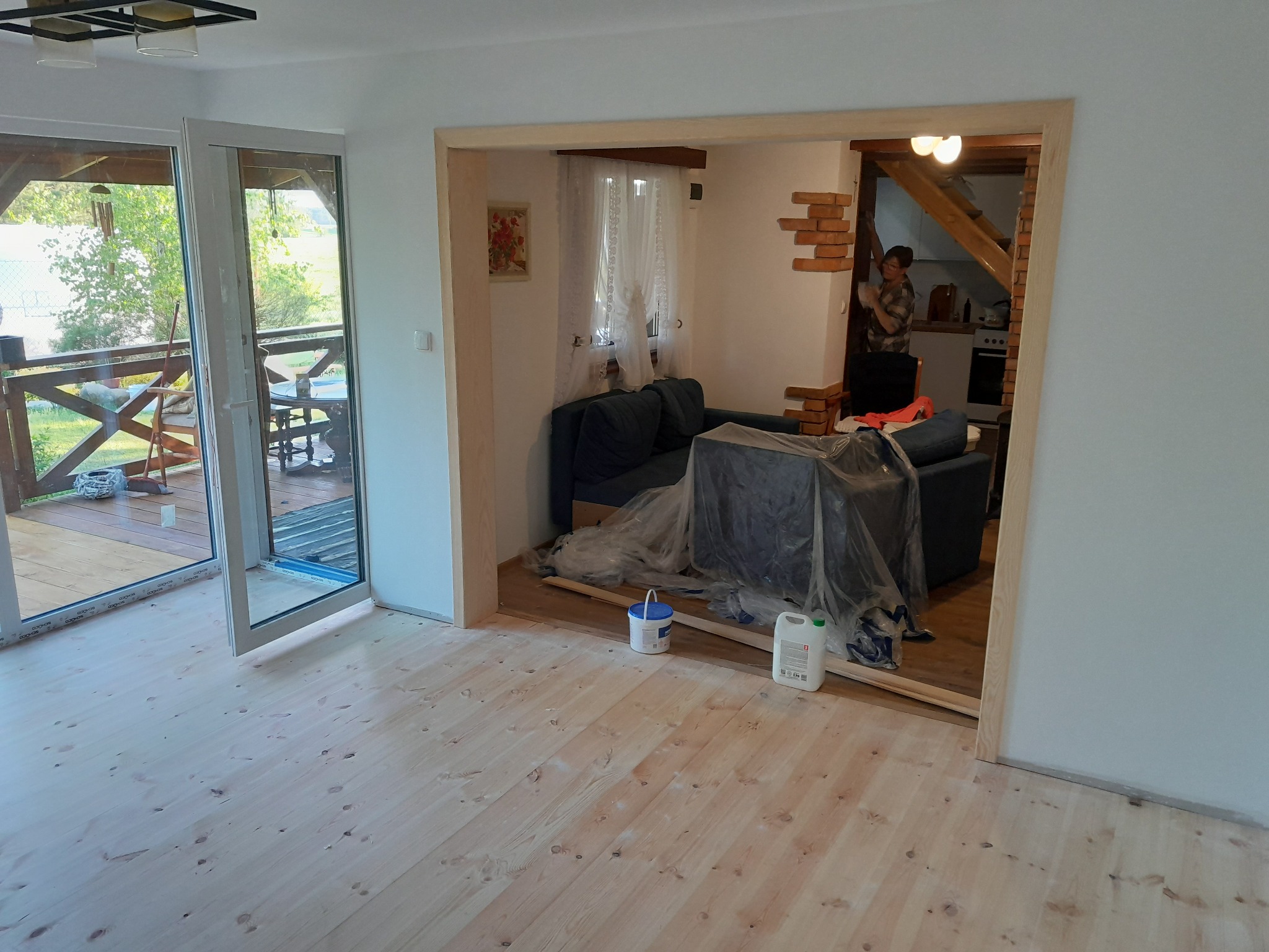 Jasna, drewniana podłoga po renowacji w przestronnym wnętrzu z widokiem na taras. Częściowo zakryte meble sugerują trwające prace wykończeniowe.