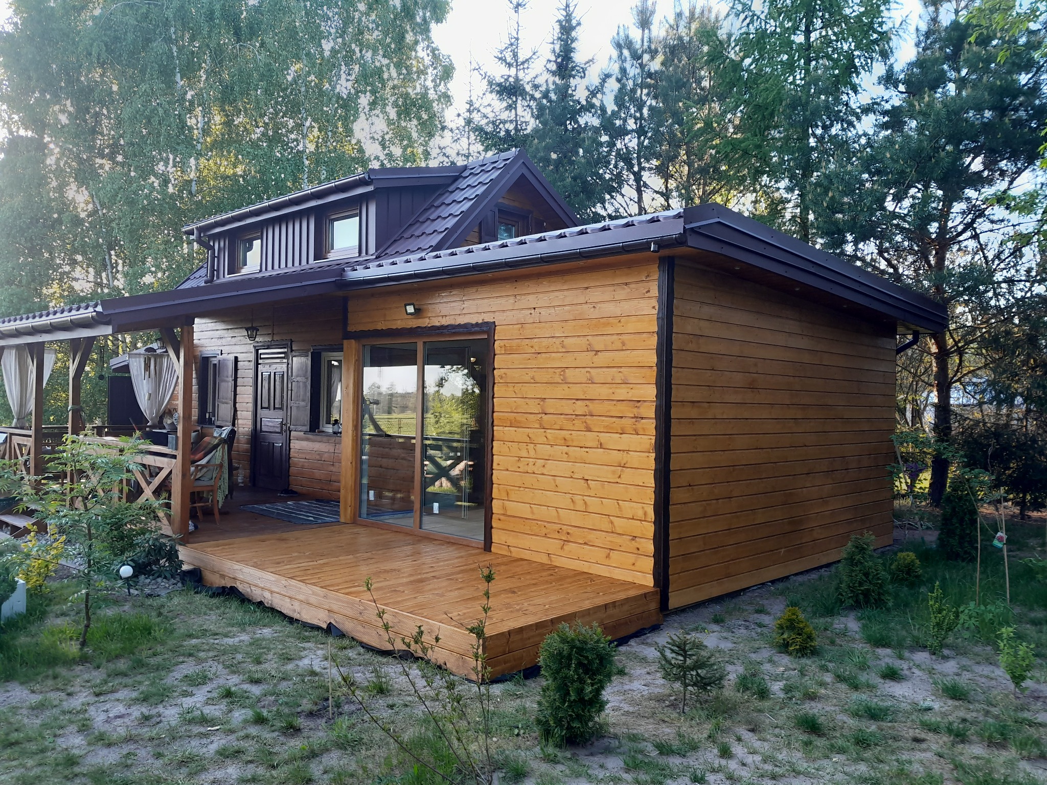 Drewniany dom z tarasem w otoczeniu zieleni. Widoczne deski tarasowe i elewacja z jasnego drewna. Nowoczesna architektura z dużymi przeszkleniami.