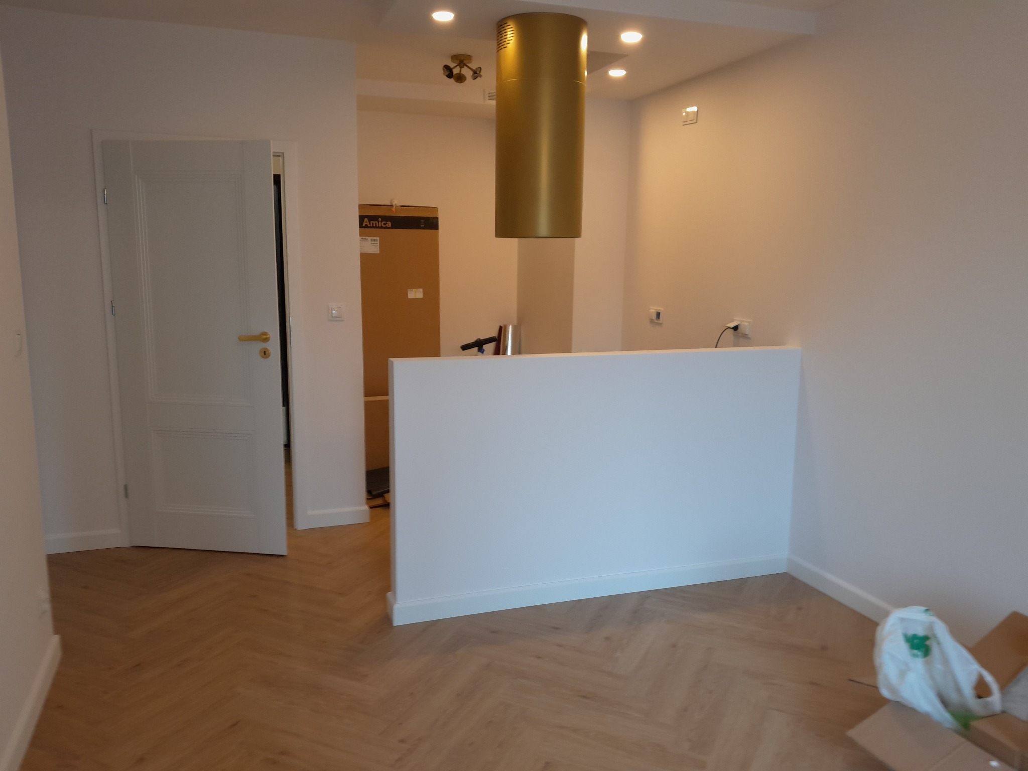 Jasne wnętrze po remoncie: parkiet w jodełkę, białe ściany, otwarta przestrzeń z elementami kuchni i złotym okapem. Widoczne detale wykończeniowe.